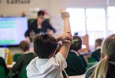 A primary school pupil with his hand in the air in front of his teacher